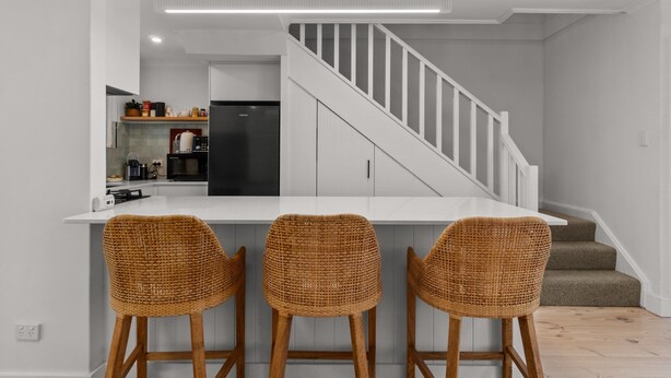 A modern kitchen with a white island, three wicker bar stools, and a staircase leading to a second floor. - Stylish 2-bed Townhouse in Leafy Hyde Park (Adelaide)