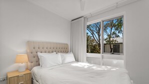A bedroom with a tufted headboard, a bedside table with a lamp, and a window with a view of trees. - Stylish 2-bed Townhouse in Leafy Hyde Park (Adelaide)