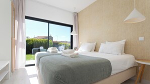 Ein modernes Schlafzimmer mit einem großen Bett, einem Nachttisch, einer Pendelleuchte und Blick auf einen Garten durch ein großes Fenster.