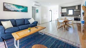 Ein modernes Wohnzimmer mit einem blauen Sofa, einem Holztisch und einem bunten Teppich. Eine Küchenzeile mit weißen Schränken und ein Esstisch mit Stühlen ist ebenfalls vorhanden.