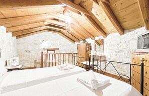 Quarto rústico com vigas de madeira, paredes de pedra, duas camas com toalhas, um armário de madeira e uma pequena mesa com abajur.