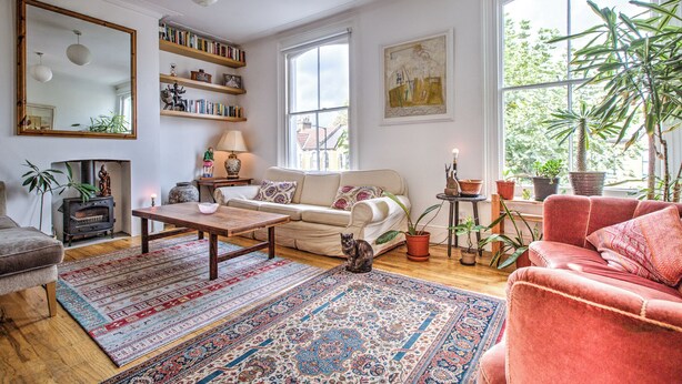 A cozy living room with a sofa, a coffee table, and a fireplace. There are bookshelves, a mirror, and a window with a view of greenery. - Charming Three Floor Retreat in Lower Clapton with Friendly Cats and Nature (London)