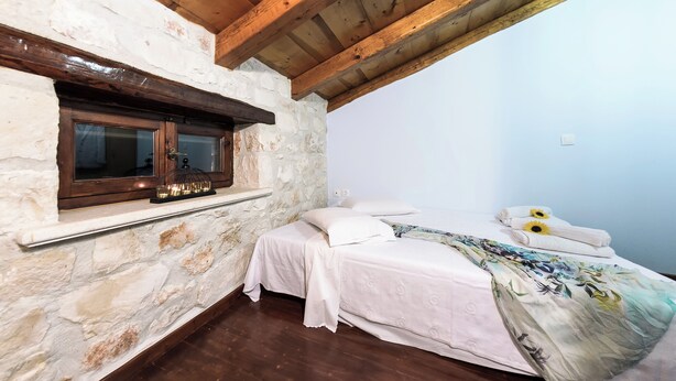 Ein Schlafzimmer mit Steinwänden, Holzbalken, einem Fenster mit Holzrahmen, einem Bett mit weißer Bettwäsche und einer dekorativen Lampe.