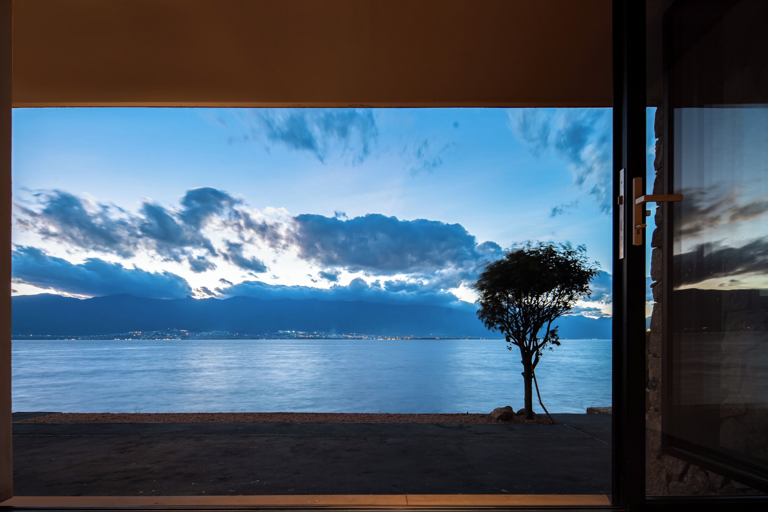 A view from inside a room with a large window overlooking a calm body of water, a tree, and distant mountains.