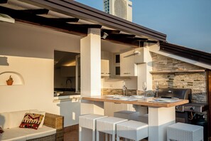 An outdoor dining area with a kitchen in the background, white wicker furniture, and a stone wall. - Stunning rooftop in Garda! (Garda)