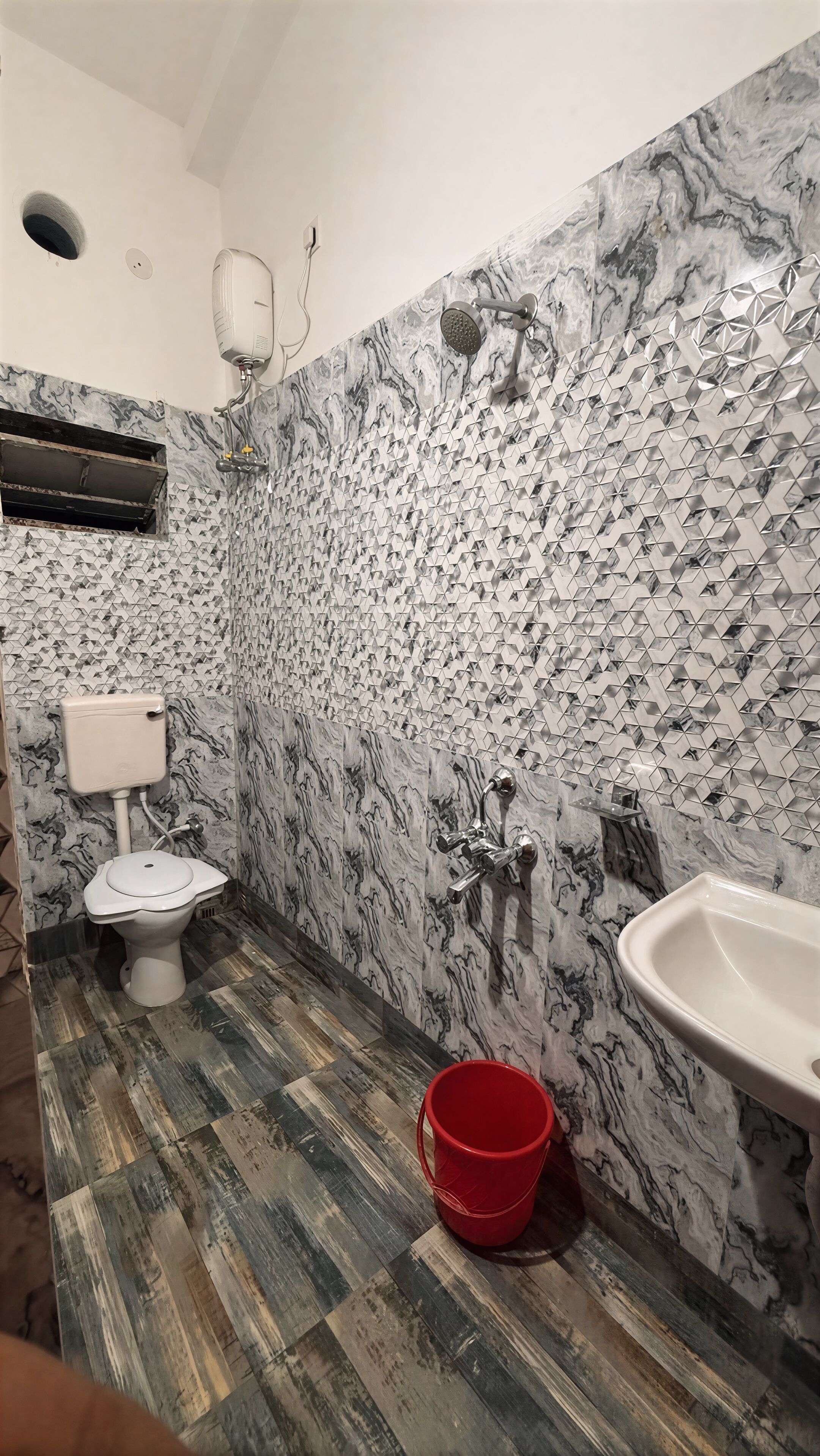 Casa de banho com revestimento em mármore, sanita, lavatório e um balde vermelho no chão.
