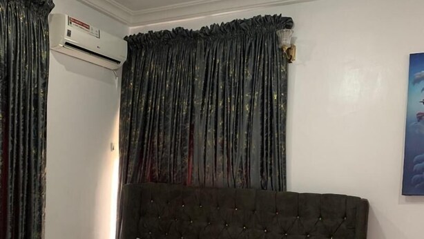 A bedroom with a tufted headboard, white bedding, a bedside table, and an air conditioning unit. - ELLEX SLEEVES APARTMENT  (Ibadan)