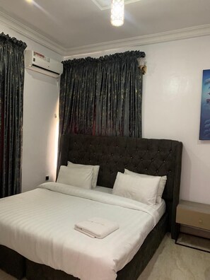 A bedroom with a tufted headboard, white bedding, a bedside table, and an air conditioning unit. - ELLEX SLEEVES APARTMENT (Ibadan)