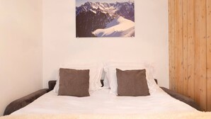 Un dormitorio con una cama, dos almohadas, una colcha blanca y un cuadro en la pared con un paisaje invernal de montañas nevadas.