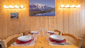 Una mesa de comedor de madera dispuesta para cuatro personas, con platos blancos, servilletas rojas y vasos transparentes. Dos lámparas de pared iluminan el ambiente.