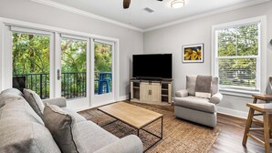 A living room with a sofa, a coffee table, a TV, and a chair. There are large windows with a view of greenery outside. - 2BR near Gulf Place w/ Pool, Grill + Free Tickets: Dolphin Cruise & More! (Santa Rosa Beach)