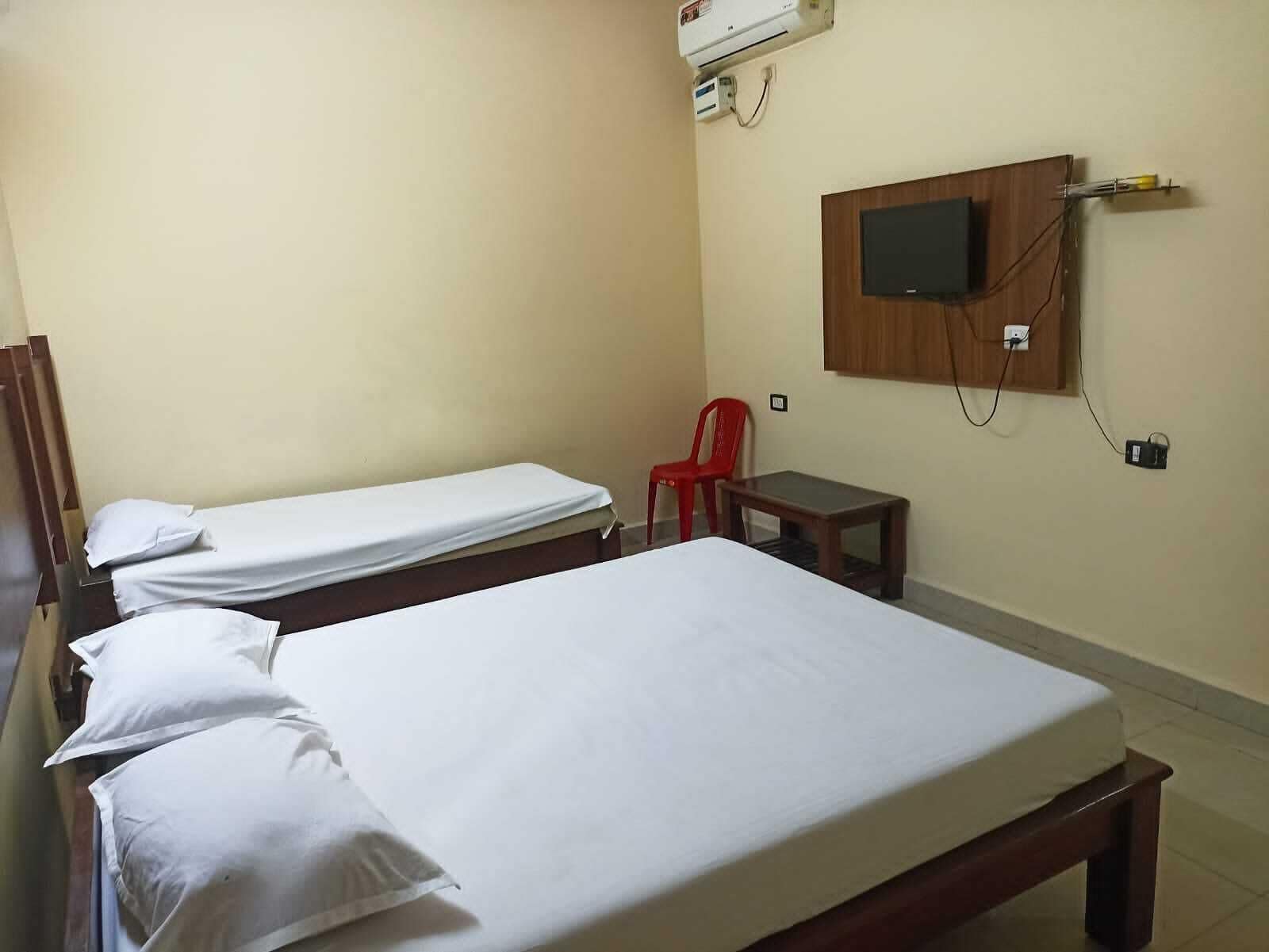 A hotel room with two single beds, a small table, a red chair, and a mounted TV.