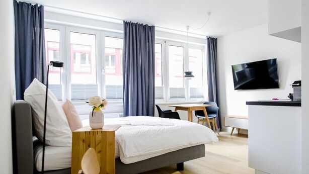 A modern hotel room with a bed, a desk, a chair, a TV, and large windows with curtains. - Louis and Louise Apartments & Lofts  (Bremen)