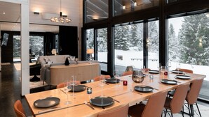 A modern dining area with a long table set for a meal, large windows offering a snowy forest view, and a cozy seating area with a sofa and lamp.