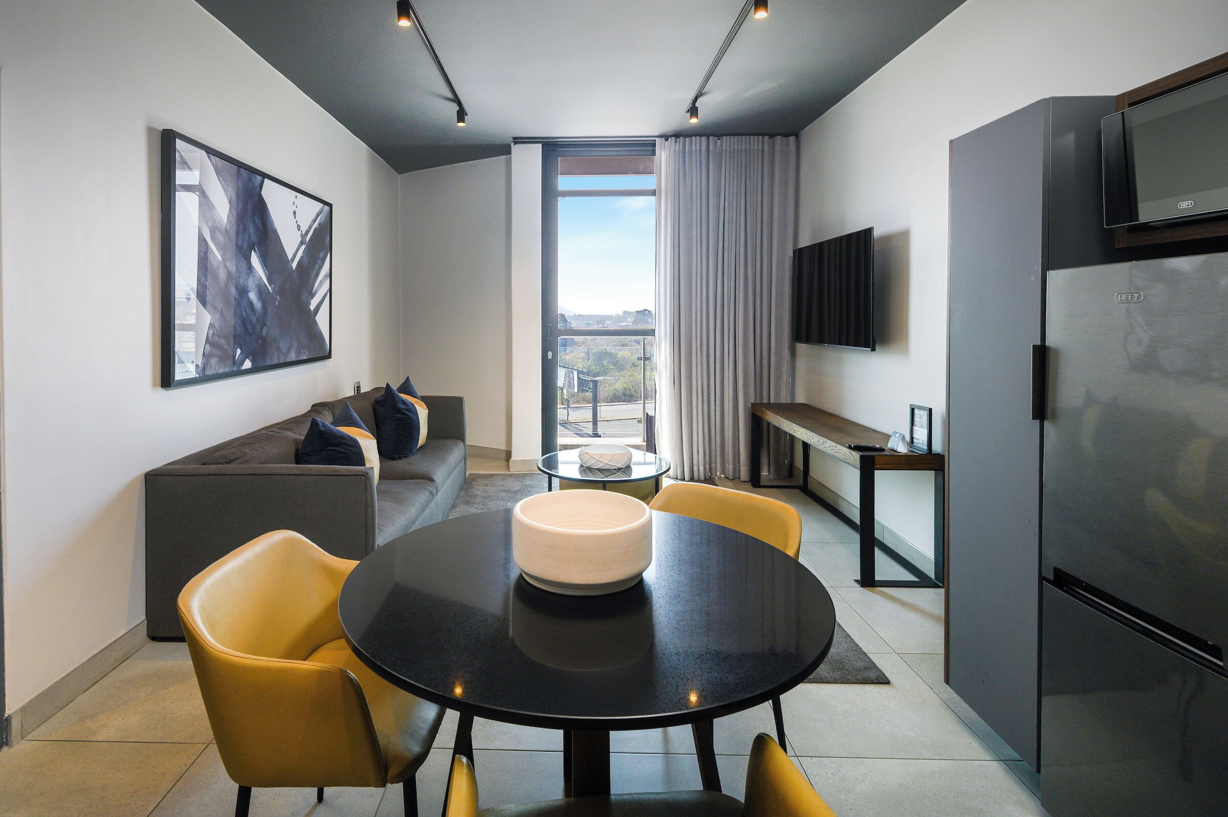 A modern hotel room with a sofa, a dining table with chairs, a flat-screen TV, and a refrigerator.