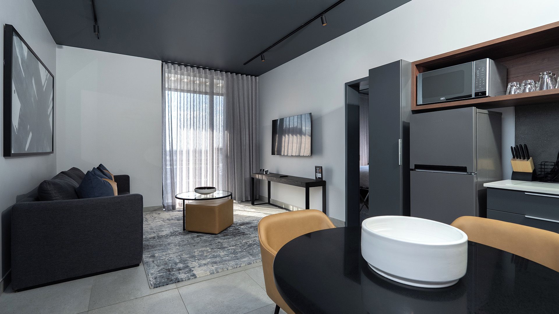 A modern hotel room with a grey sofa, a round dining table with a white bowl, a small side table, a desk with a chair, a flat-screen TV, and a built-in kitchen area with a microwave and refrigerator.