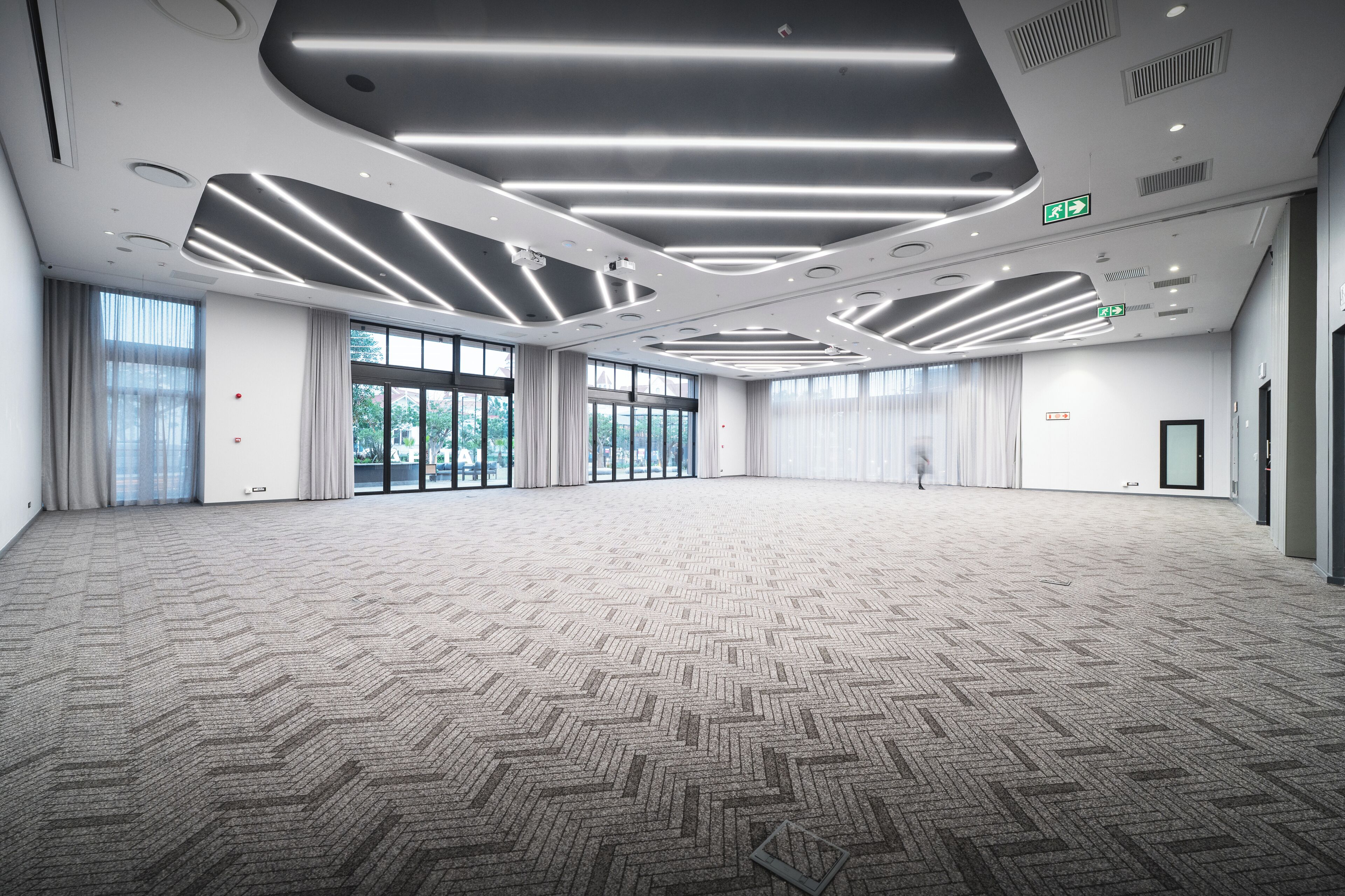 Una sala de conferencias amplia, bien iluminada, con un diseño moderno en el techo, ventanas grandes y una alfombra estampada.