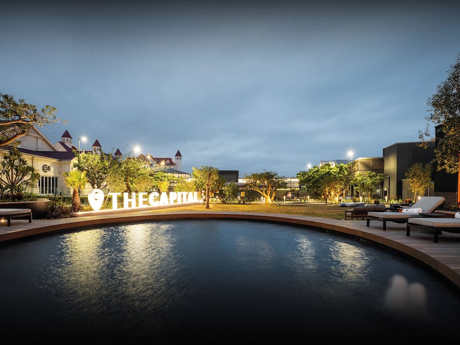 Una zona de piscina con sillones de descanso, un cartel que dice 'THE CAPITAL' y edificios al fondo.
