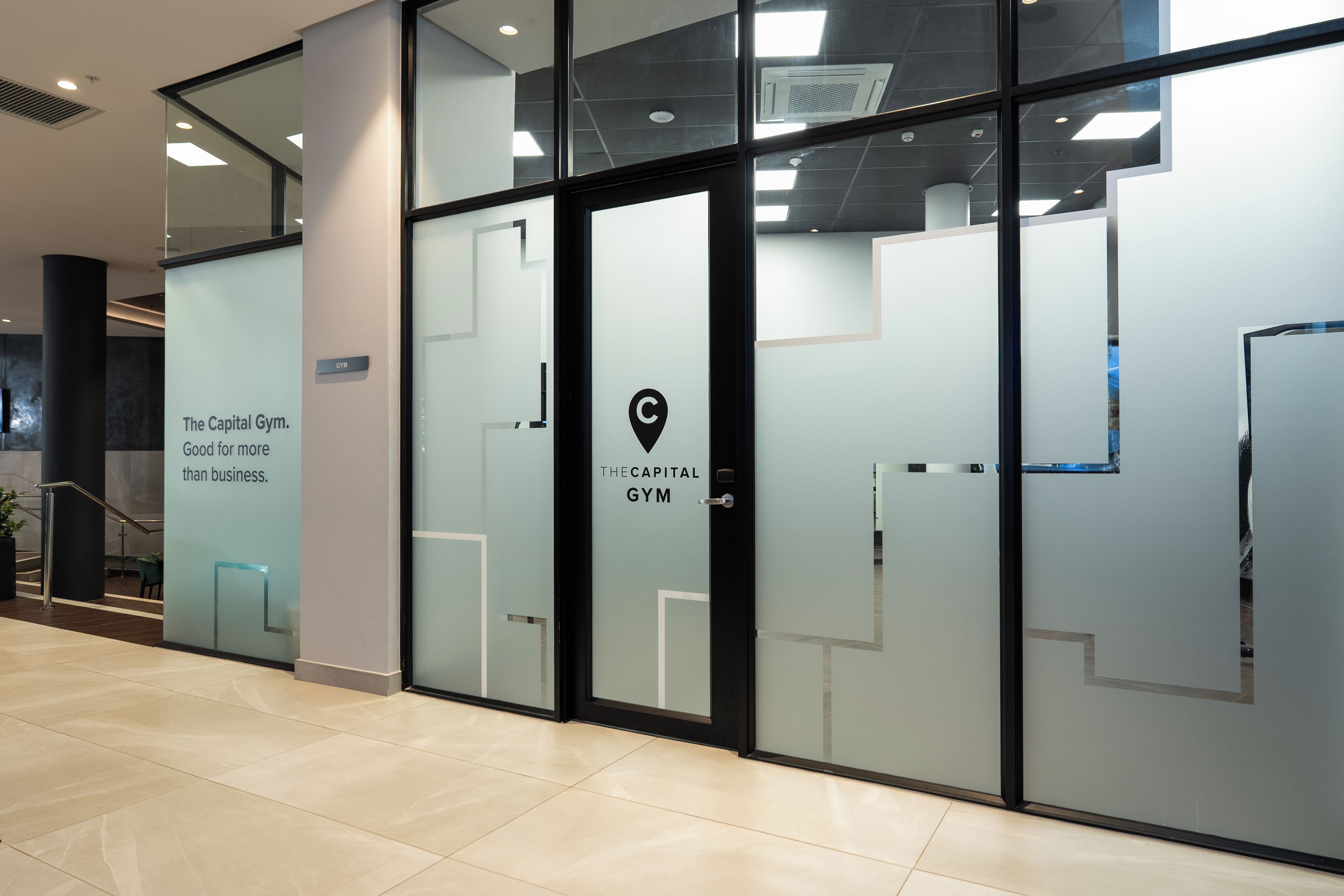A modern building entrance with glass doors, a sign for 'The Capital Gym,' and a logo indicating a location.
