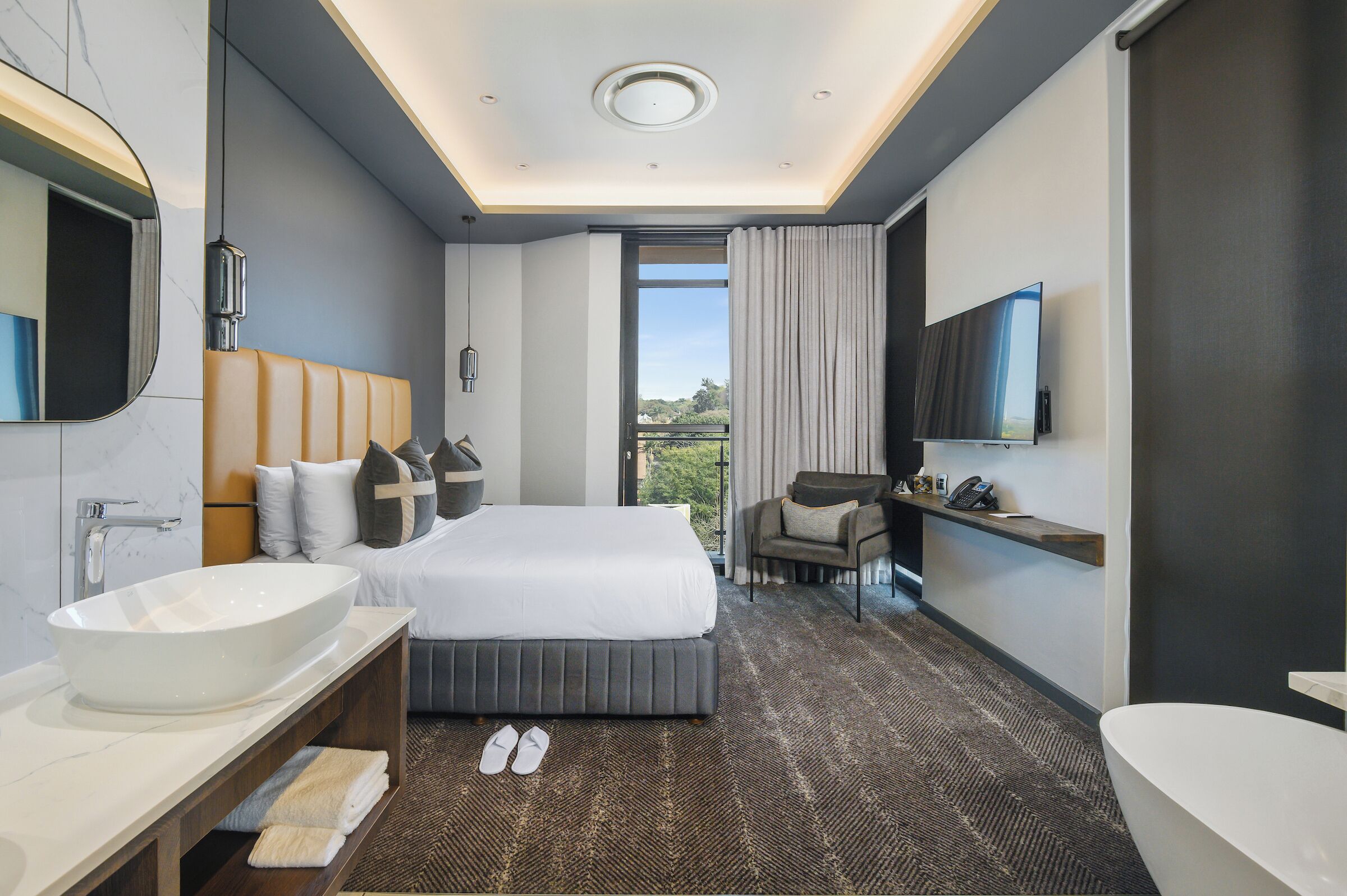 A modern hotel room with a large bed, a bathroom with a bathtub, a vanity with a large mirror, and a television mounted on the wall.