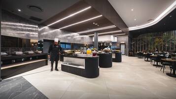 A modern restaurant with a chef, a buffet counter, and a seating area with tables and chairs.