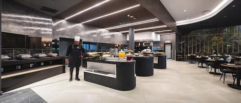 Un restaurante moderno con un chef, una mesa de buffet y un área de comedor con mesas y sillas.