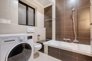 A modern bathroom with a washing machine, toilet, and a bathtub with a showerhead. - Spacious waterfront 1-bedroom apartment GSPI1-1 (Birgu)