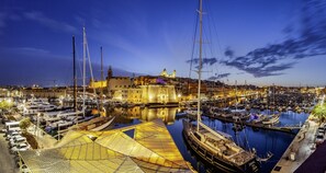 Un port de plaisance avec des bateaux amarrés, un grand voilier et un bâtiment historique surmonté d’un dôme, le tout sur fond de paysage.