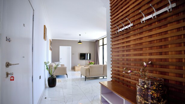 A modern living room with a sofa, a coffee table, and a TV. There is a wooden wall with hooks and a vase with flowers. - Le Basilic Addis Hotel Apartment & Spa (Addis Ababa)