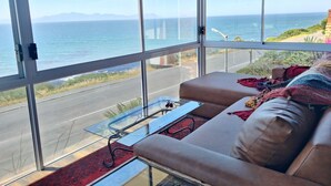 A room with a large window offering a view of the ocean and a road. There is a sofa with a blanket and a glass table with a pair of sunglasses on it. - Boho Ocean-View Villa: Pool & Walk to Beach (Murdock Valley)