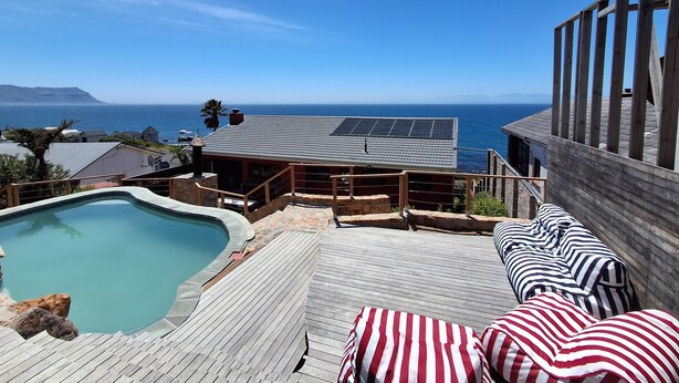 A pool area with a view of the ocean, a wooden deck, and a building with solar panels. - Boho Ocean-View Villa: Pool & Walk to Beach (Murdock Valley)