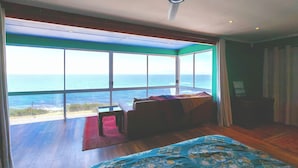 A bedroom with a large window overlooking the ocean, a ceiling fan, and a bed with a patterned bedspread. - Boho Ocean-View Villa: Pool & Walk to Beach (Murdock Valley)