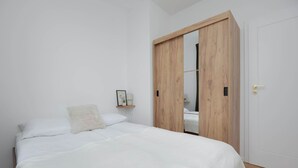 A bedroom with a bed, a wooden wardrobe, a white door, and a small shelf with a picture and a plant.