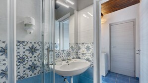 Un baño con lavamanos blanco, azulejos azules, cabina de ducha de vidrio y una puerta que da a otra habitación.