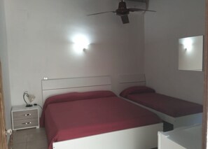 Une chambre avec deux lits simples, une table de chevet avec une lampe, un ventilateur de plafond et un luminaire fixé au mur.