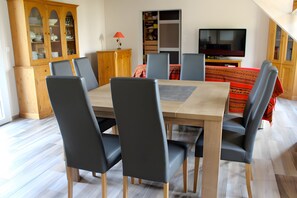 A dining area with a wooden table and grey chairs, a cabinet with glass doors, and a television in the background.