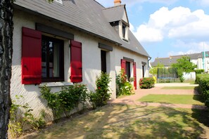 A house with red shutters, a white exterior, and a garden with bushes and a lawn.
