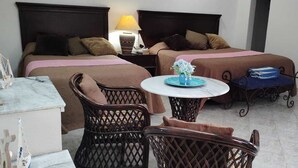 Habitación de hotel con dos camas, una mesita con un florero, sillas de mimbre y una puerta de madera.