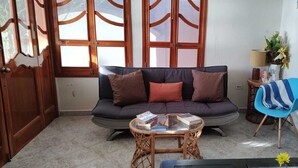 Una sala de estar con un sofá oscuro, una mesa de centro redonda de mimbre y una silla azul.