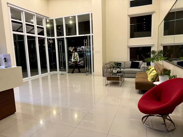 A spacious lobby with a modern design, featuring a seating area with a red chair, a sofa, and a coffee table. There is a glass door leading to the outside, and a person in a uniform is visible near the door.
