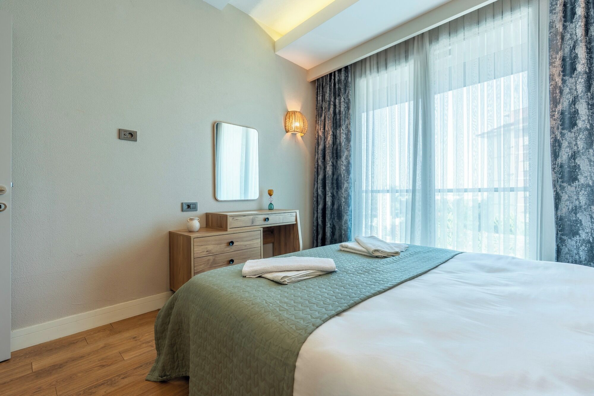 Habitación de hotel con cama, mesita de noche, espejo, luz empotrada en la pared y ventana con cortinas.