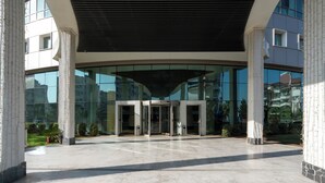 A modern building entrance with glass doors, a covered walkway, and a reflection of the building in the glass. - Maxihomes Snitch (Antalya)