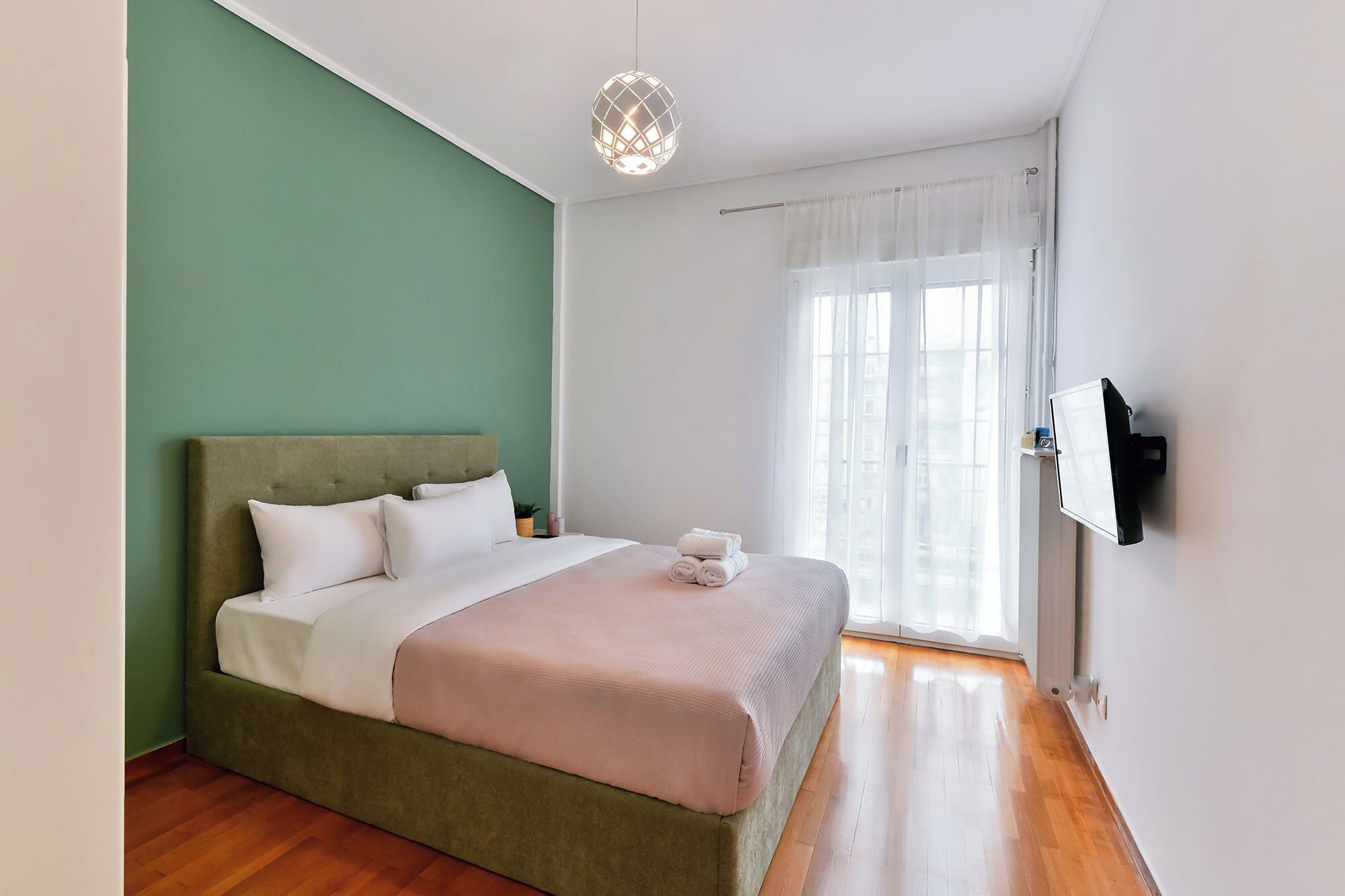 Een slaapkamer met een bed, nachtkastje, een aan de muur bevestigde televisie en een raam met gordijnen.