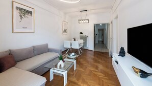 A modern living room with a sofa, coffee table, and TV. There is a dining area with a table and chairs. The room has wooden flooring and white walls. - Porto Sea View Apartments - Ermou 61  (Thessaloniki)