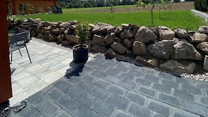 A patio with stone tiles, a stone wall, a small potted plant, and a view of a green field and trees under a clear sky. - Apartment 'Ferienwohnung Sußebach mit 80 qm' (Vöhringen)