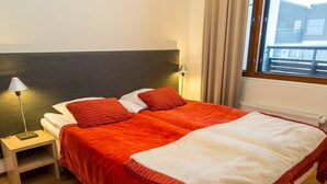 Un lit double avec une couverture rouge et blanche, deux oreillers rouges, une table de chevet avec une lampe et une fenêtre avec des rideaux.