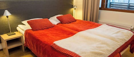 A double bed with red and white bedding, two bedside lamps, a wooden nightstand, and a window with curtains.