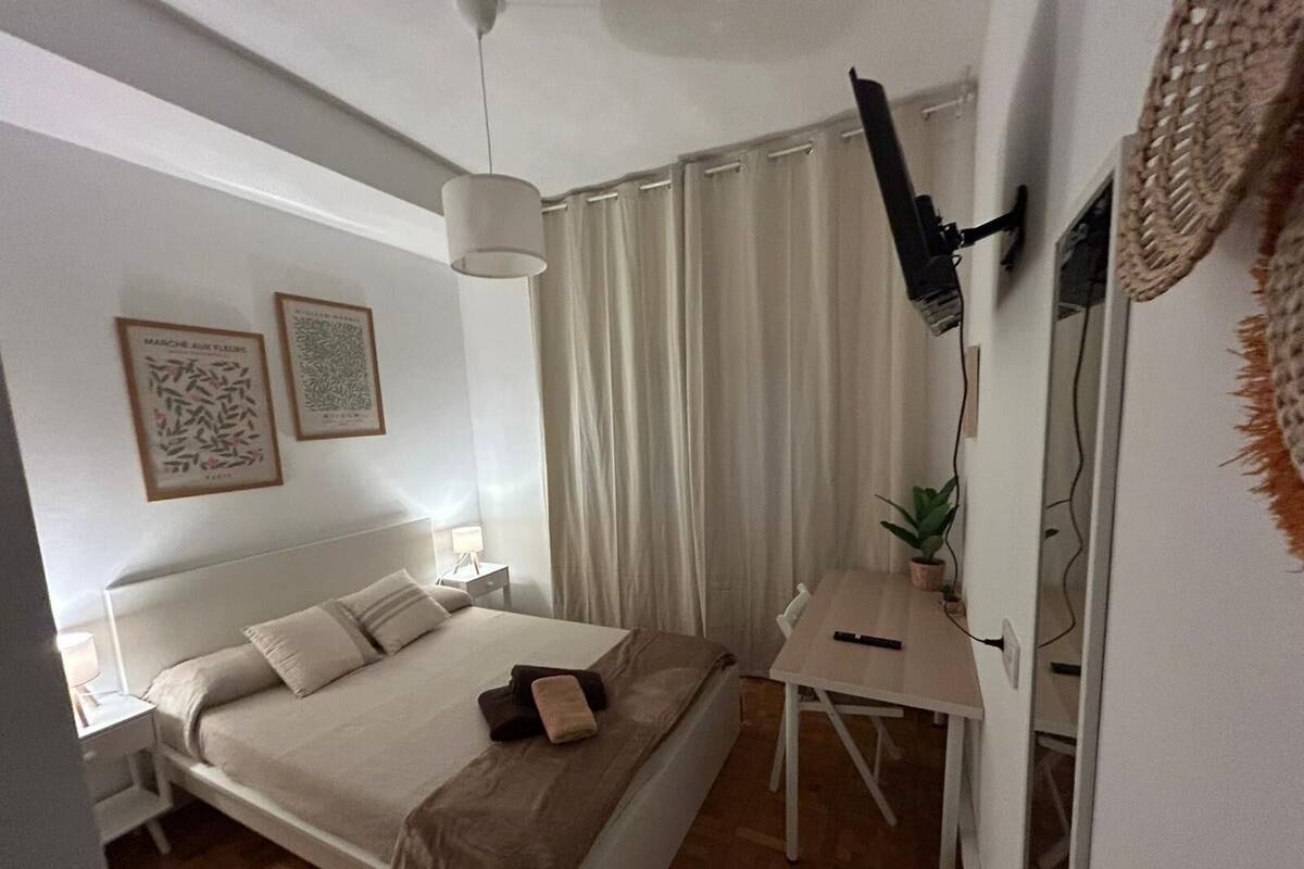 Ein Schlafzimmer mit einem Bett, Nachttischen, einem Schreibtisch, einem Sessel, einer Pendelleuchte, einer Zimmerpflanze und einem Vorhang.