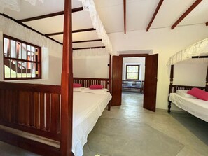 Una habitación con dos camas de madera, cada una con colcha blanca y almohadas rojas. El techo es de madera con vigas a la vista y el suelo de hormigón. Hay una ventana con vistas a una escalera y una puerta que conduce a otra habitación.