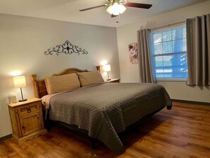 Um quarto com cama, mesas de cabeceira, ventoinha no teto, janela com cortinas e um objeto decorativo na parede.